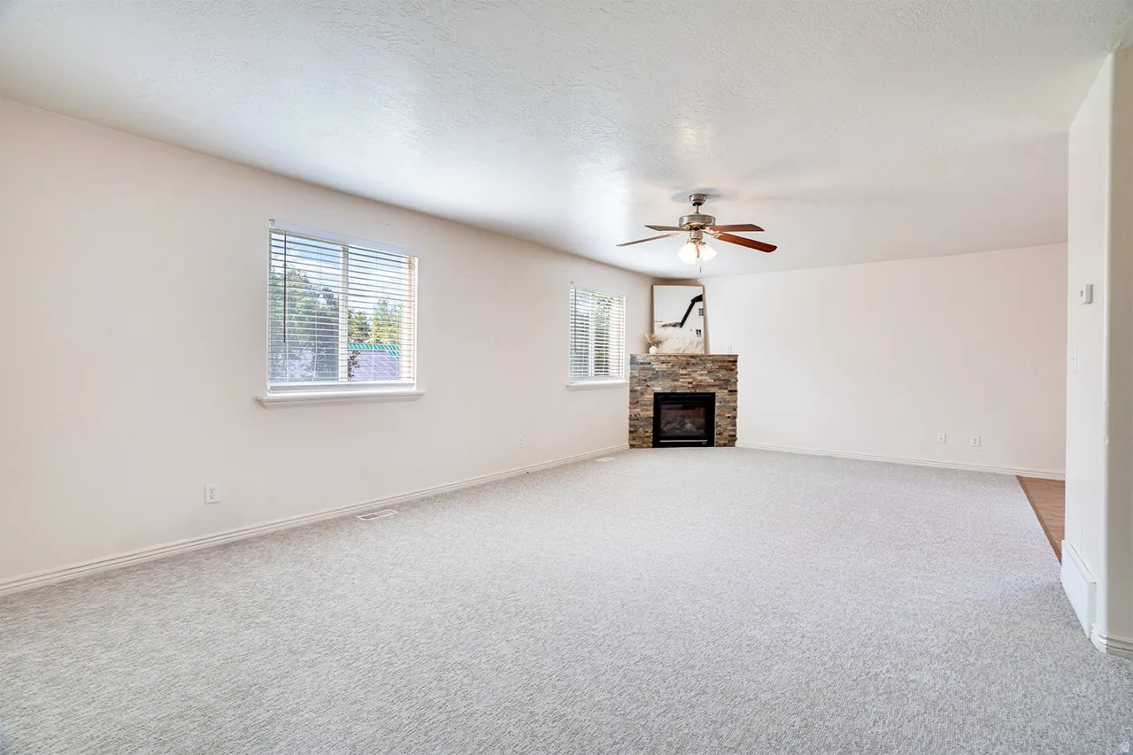 Open living area featuring light-toned carpeting, a stone-clad corner fireplace with an electric insert, and a ceiling fan