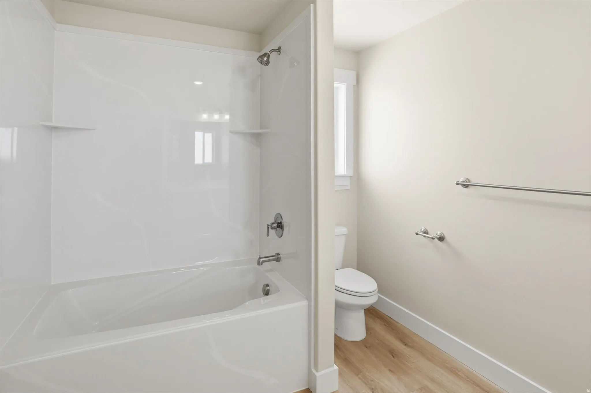 Bathroom featuring a white tub and shower combination with integrated shelving, light-toned walls, wood-finish flooring, a toilet, and brushed nickel fixtures