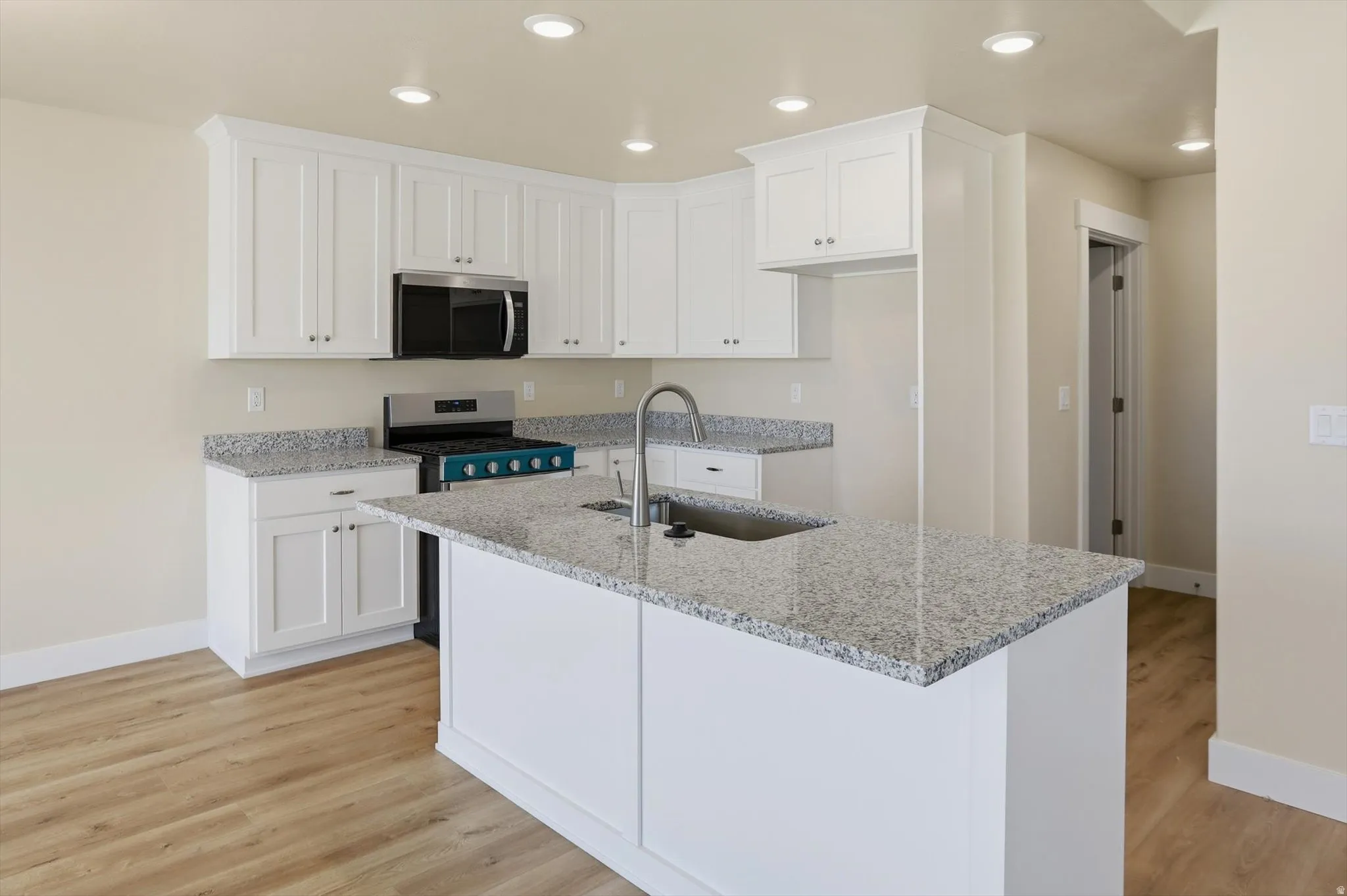 Modern kitchen featuring a central island with an undermount sink, white cabinetry, speckled countertops, stainless steel appliances, and wood-finish flooring