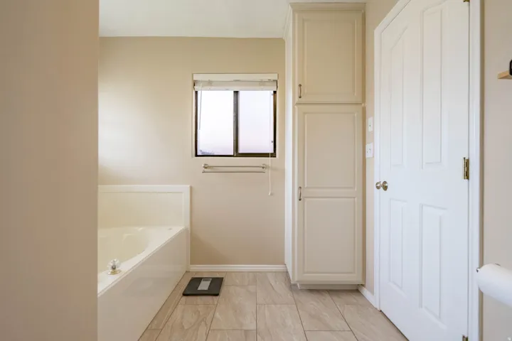 Built-in bathtub with a corner design, tile flooring, window with privacy blind, and integrated cabinetry for linen