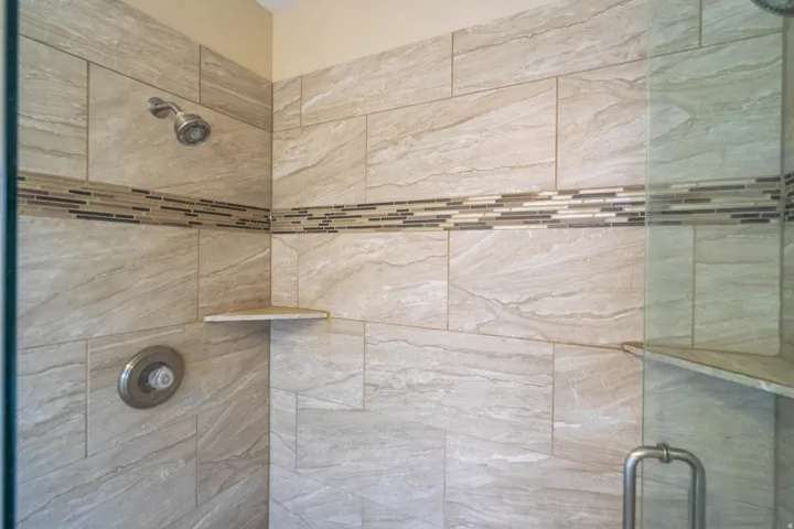 Primary Shower featuring large format tiled walls, a decorative mosaic tile accent band, a built-in corner shelf, and a glass enclosure with a brushed metal handle