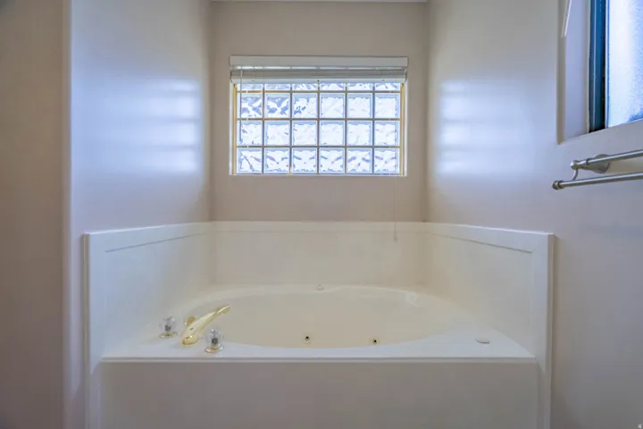 Corner-set soaking tub with a glass block window and a roller shade in primary bathroom