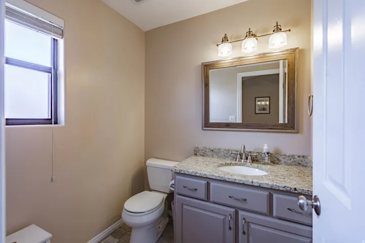 Half Bathroom featuring a single window with blinds, a toilet, and a vanity with a light-colored countertop and a framed mirror