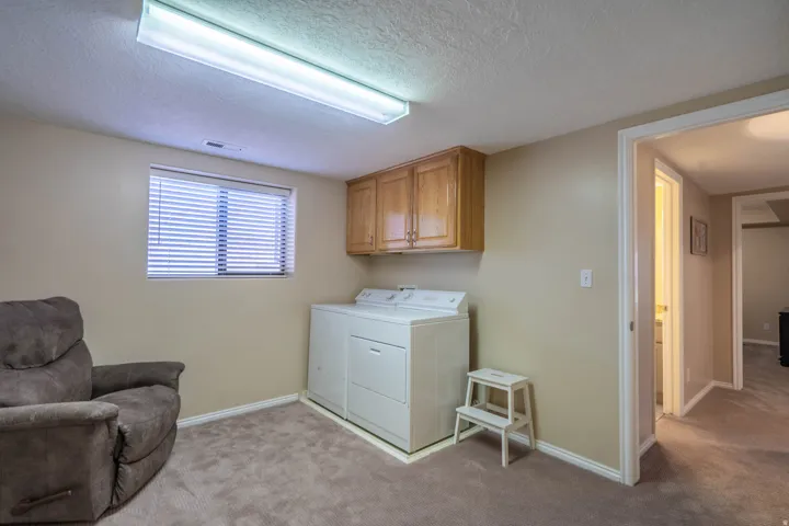 Dedicated laundry area with upper wood cabinetry, window with blinds, and neutral carpet flooring