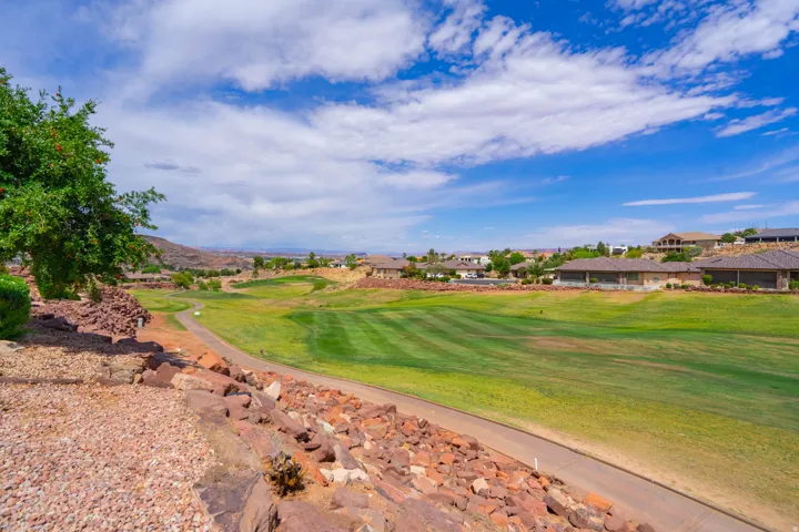 Expansive green golf course with striped fairways, surrounded by natural rocklandscaping and scattered residential properties under a clear sky as viewed from lower patio