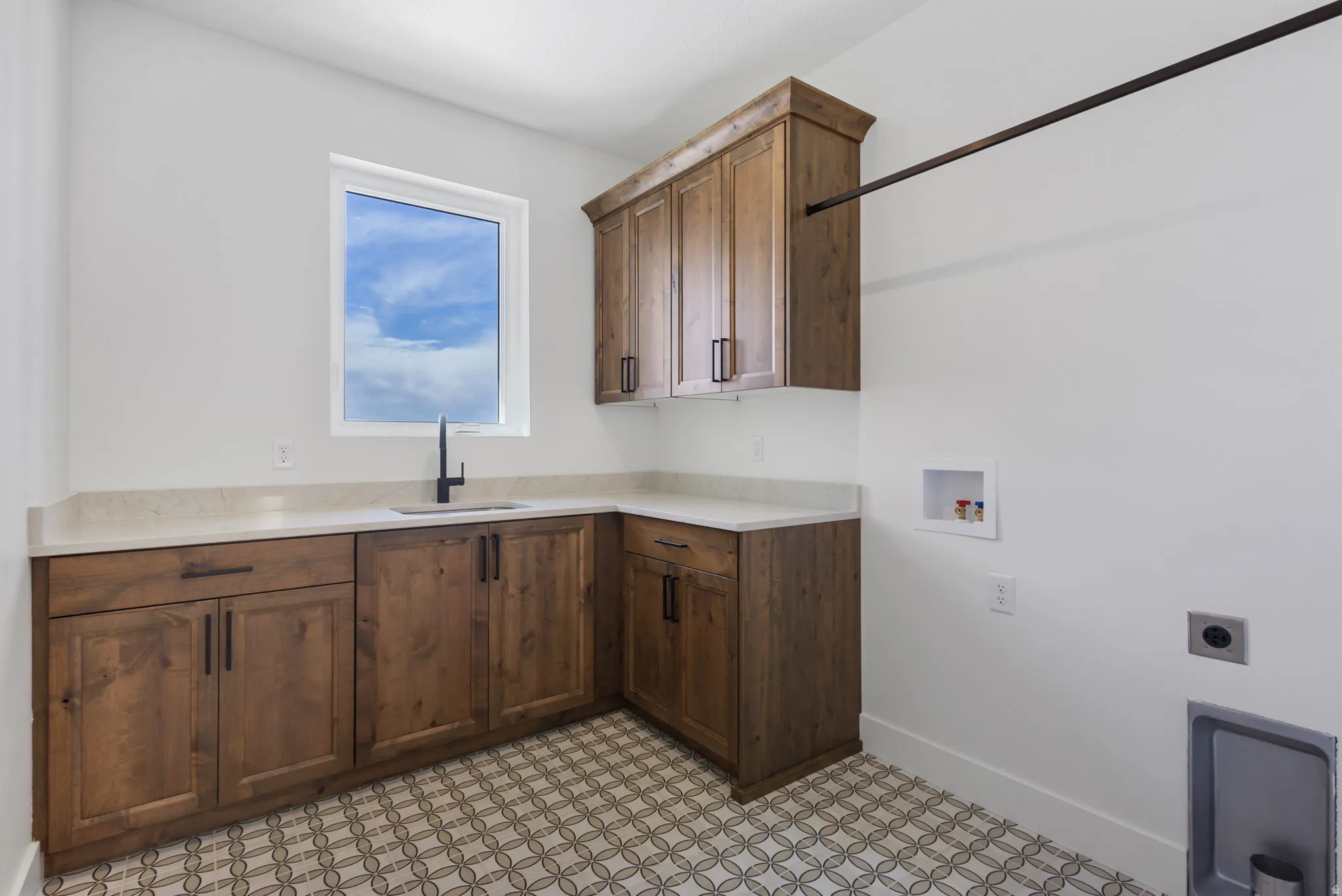 Laundry area featuring wood cabinetry, light-toned countertops, a matte black faucet, and patterned floor tiles