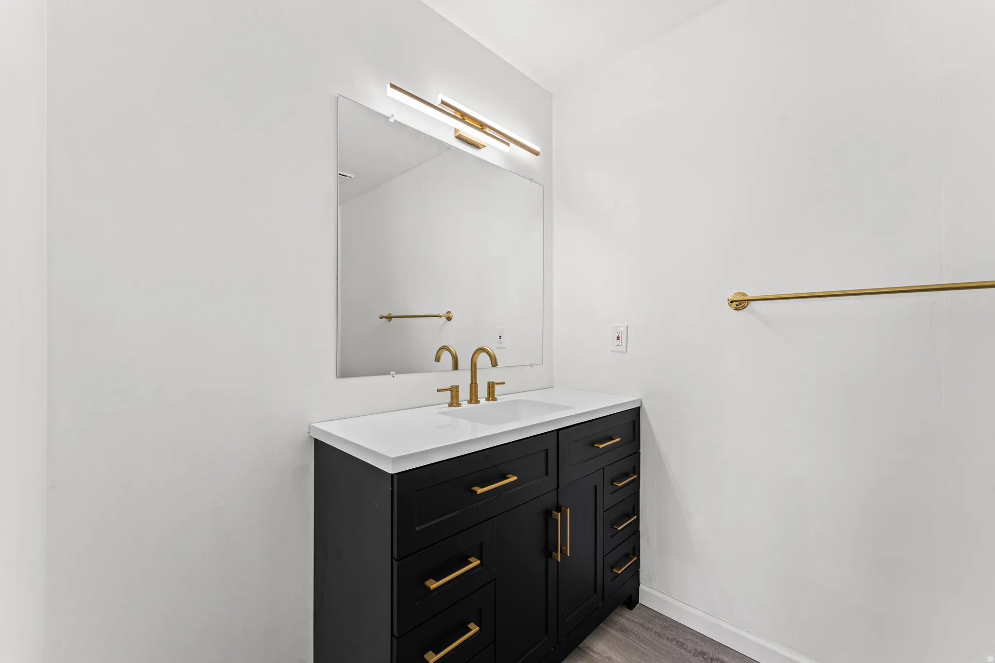 Modern bathroom vanity featuring a black cabinet with gold-finish hardware, a white countertop with an integrated sink, and a rectangular mirror with an overhead linear light fixture