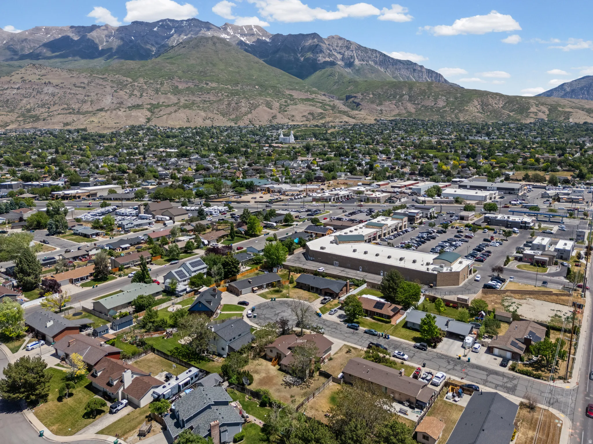 Expansive aerial view of a residential community situated at the base of prominent mountains, featuring various single-family homes, commercial buildings with parking, and established tree-lined streets
