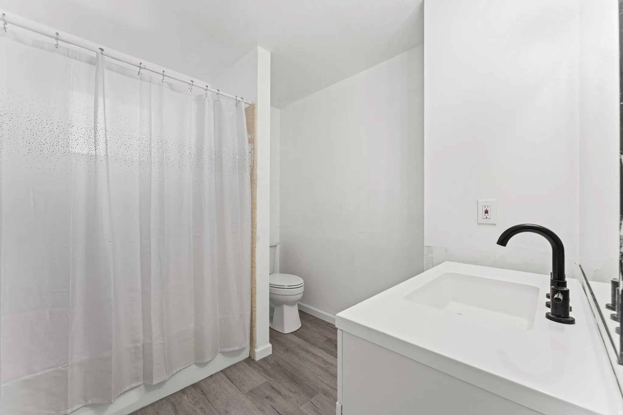 Bathroom featuring wood-finish flooring, a white vanity with an integrated sink, a black gooseneck faucet, and a shower with a white curtain