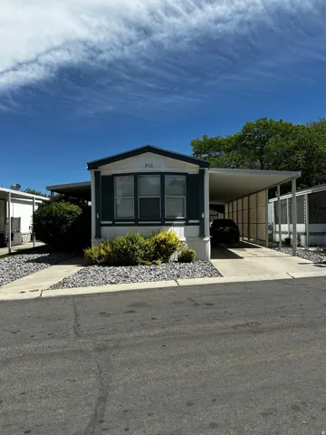 Manufactured home featuring a covered carport, pitched roofline, and an exterior with white siding and dark shutters