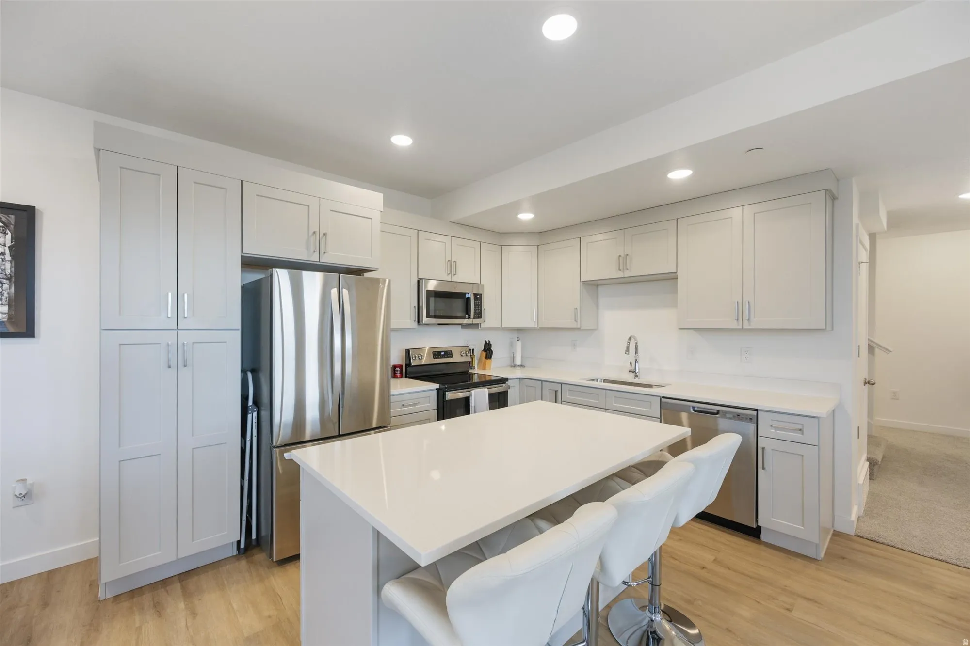 Modern kitchen featuring light gray shaker cabinetry, stainless steel appliances, a white countertop island with seating, recessed lighting, and wood-finish flooring