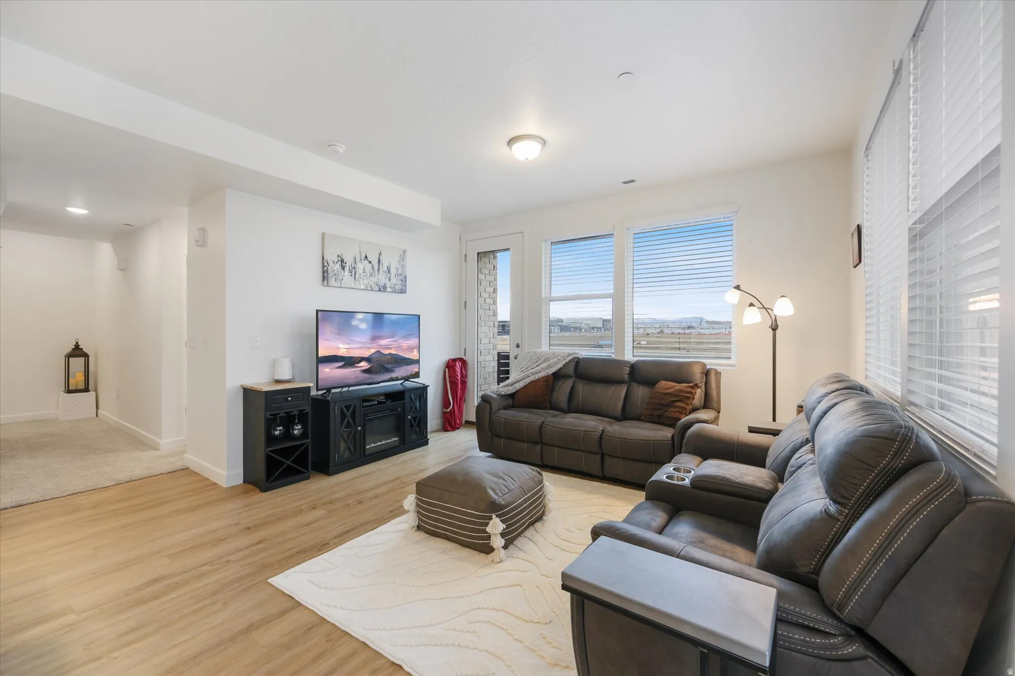 Spacious living area featuring light wood-finish flooring, white walls, and multiple windows with horizontal blinds