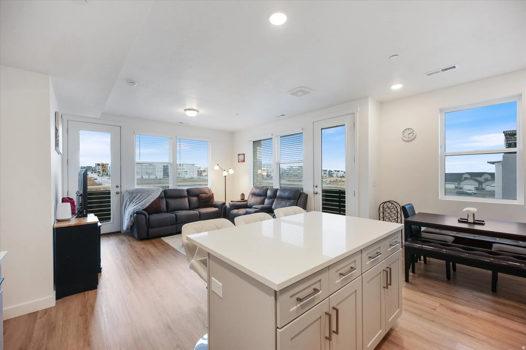 Open-concept living area featuring a kitchen island with light-colored cabinetry, wood-finish flooring, recessed lighting, and multiple windows with blinds
