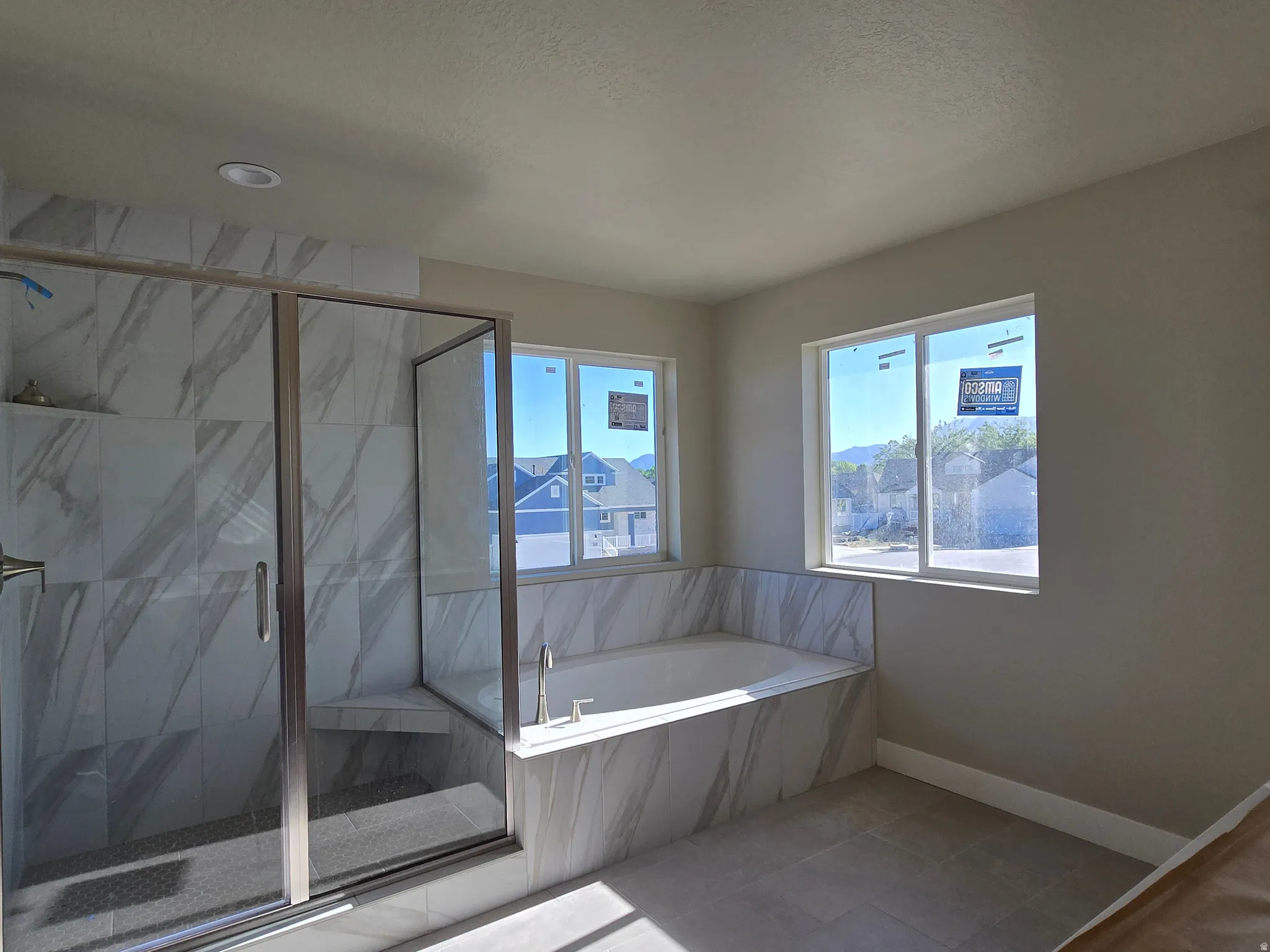 Modern bathroom featuring a spacious walk-in shower with clear glass enclosure and built-in bench, a large soaking tub with tile surround, large format tile flooring, and two expansive windows providing natural light