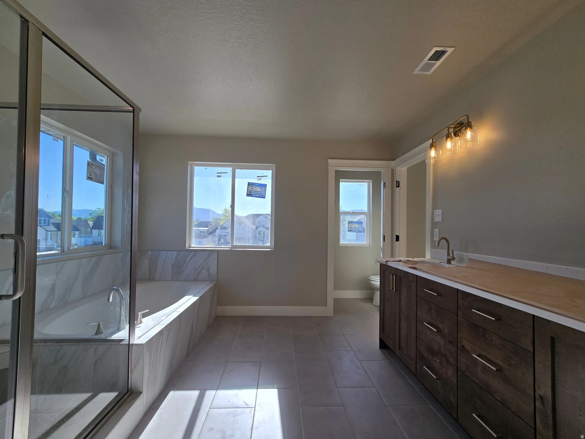 Spacious bathroom featuring a glass-enclosed shower, a large soaking tub with marble-finish tile surround, a long dark wood-finish vanity cabinet, a contemporary wall-mounted light fixture, and light-toned floor tiling