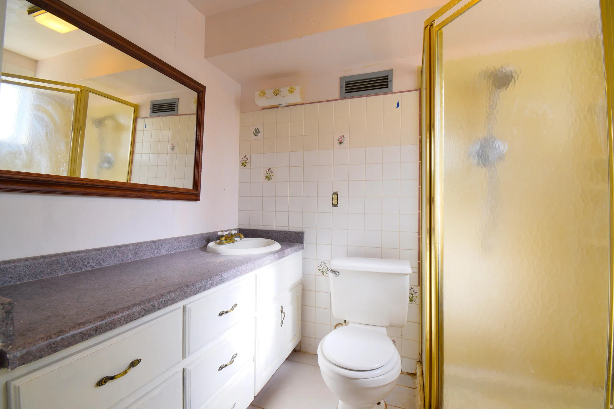Bathroom featuring a single vanity with a solid surface countertop, white cabinetry with brass-tone hardware, a framed wall mirror, a toilet, and a glass-enclosed shower with gold-tone framing
