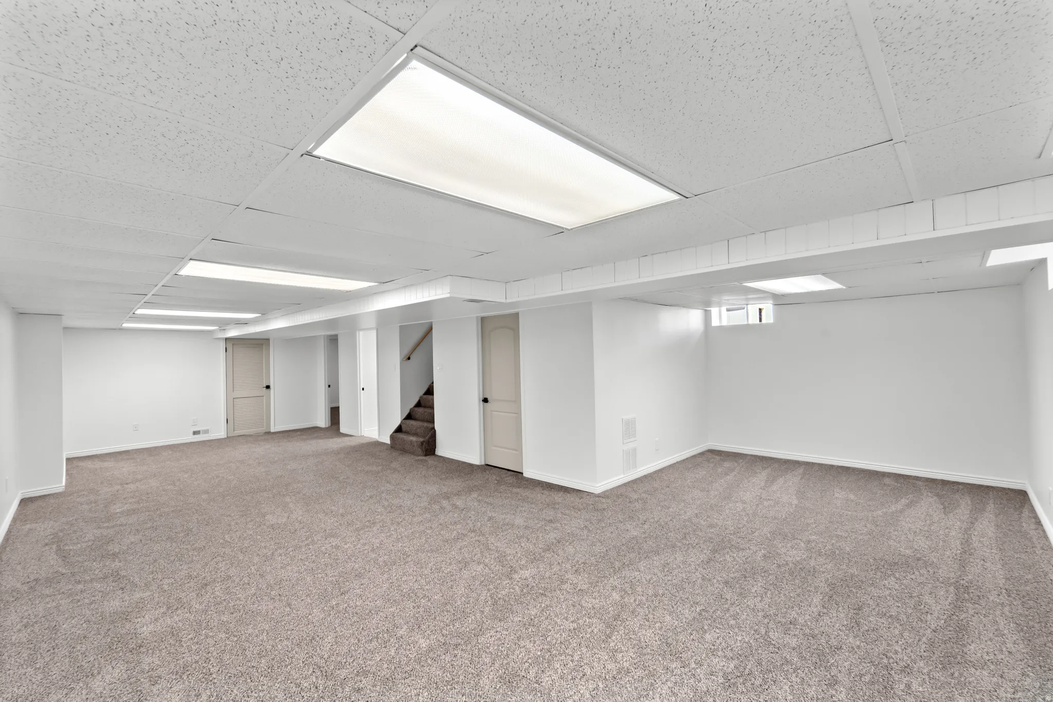 Expansive finished basement featuring neutral-toned carpeting, white painted walls, a dropped ceiling with integrated lighting, and multiple paneled doors