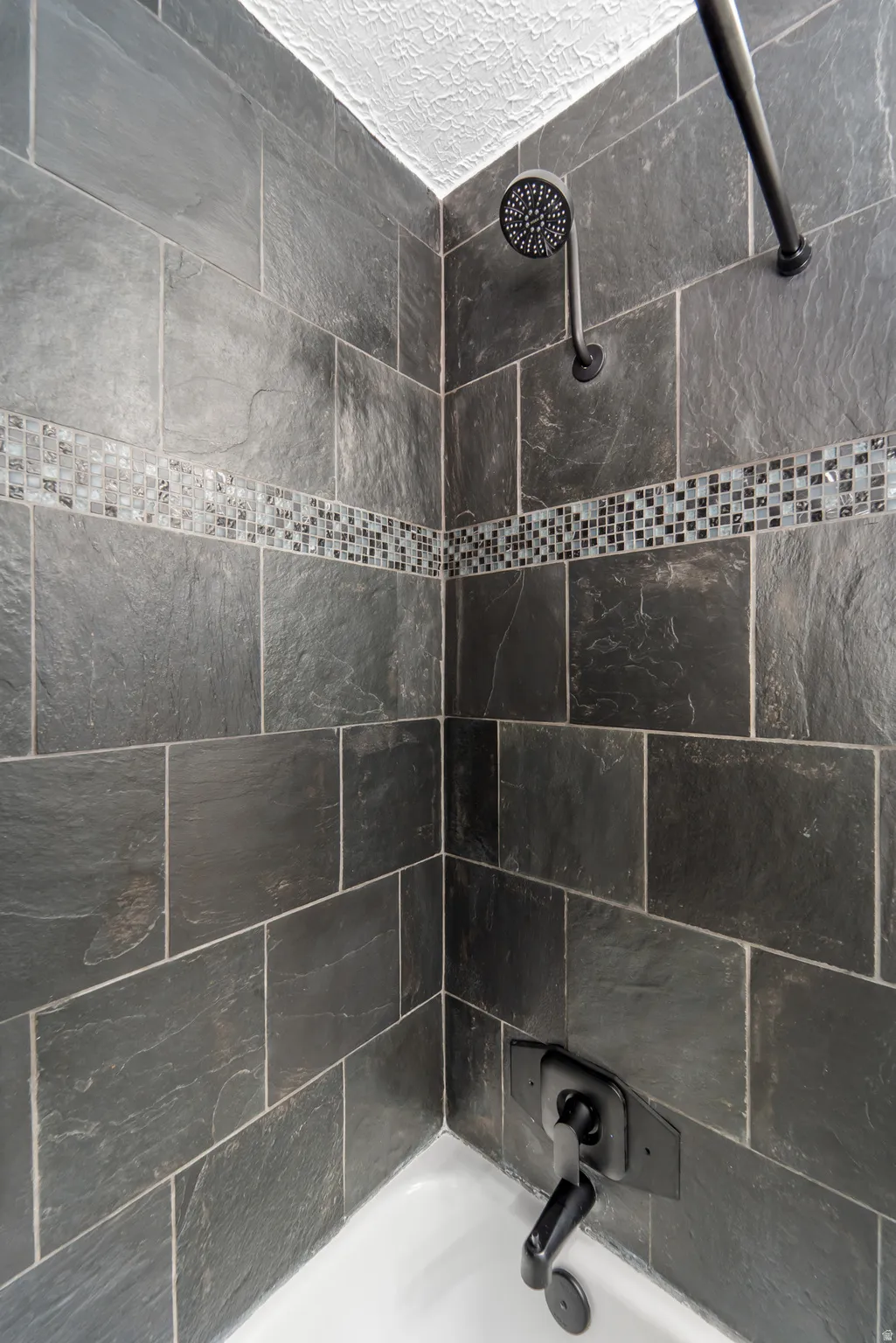 Bathtub shower combination featuring dark slate-style tile surround with mosaic accent band, a matte black shower head, and coordinating tub spout with control