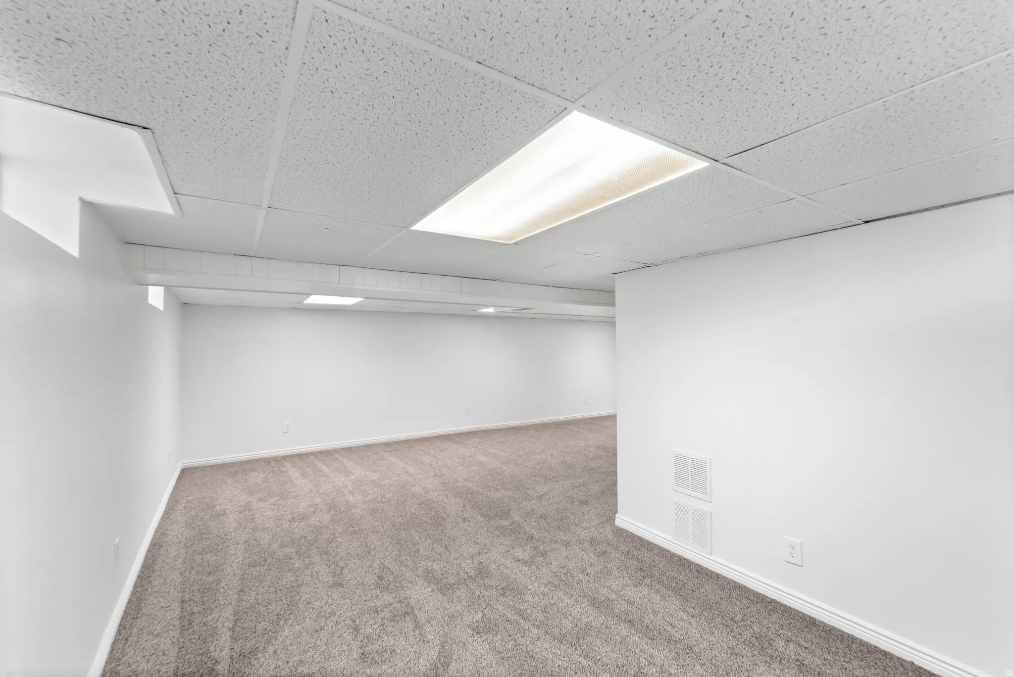 Expansive room featuring light gray carpeting, white painted walls, and a drop ceiling with integrated fluorescent lighting