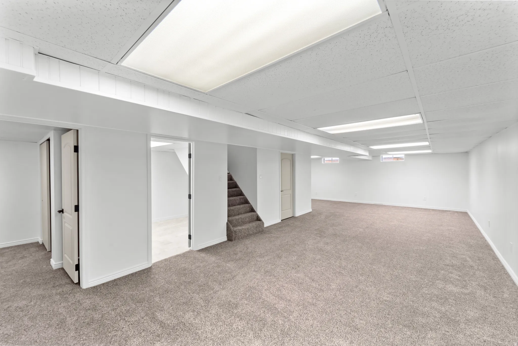 Expansive carpeted area featuring a drop ceiling with integrated lighting, white walls, and a staircase