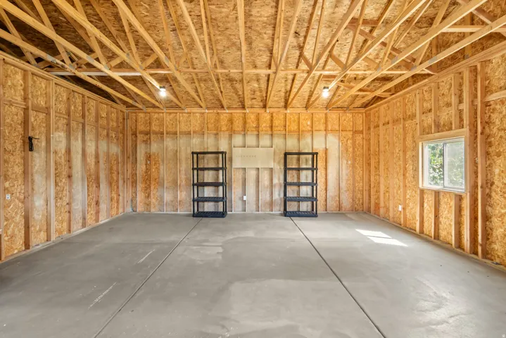 Spacious garage featuring a concrete floor, exposed wood framing, an open truss ceiling, two ceiling-mounted light fixtures, and a single window