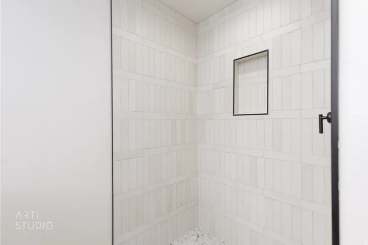 Shower enclosure featuring vertical white subway tile with horizontal accents, a recessed niche with black trim, black shower controls, and a dark pebble floor