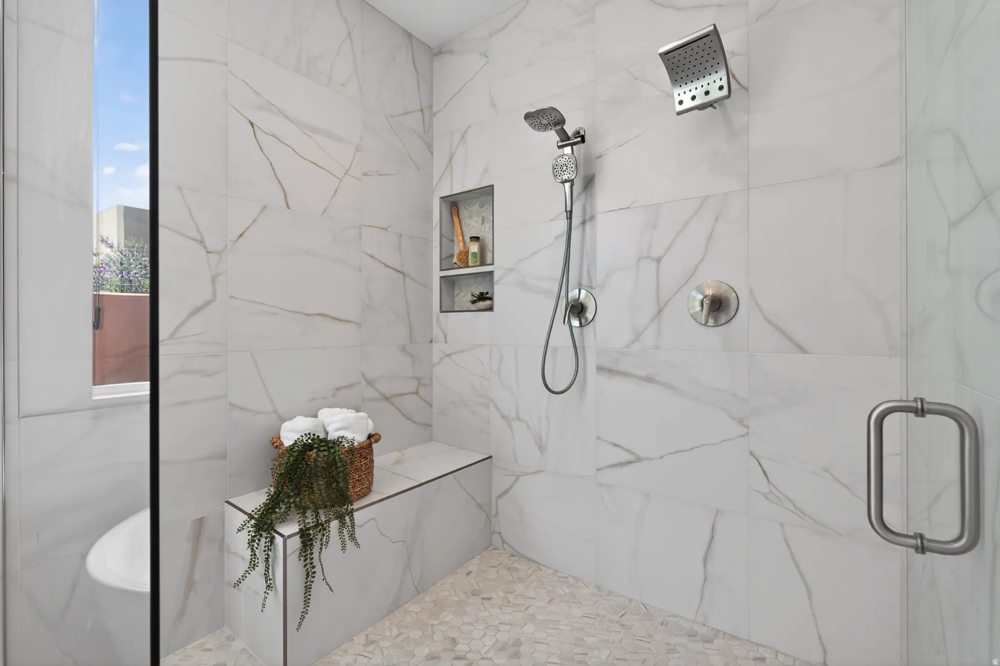 Shower with white veined tile walls, pebble floor, built-in bench, and dual shower heads