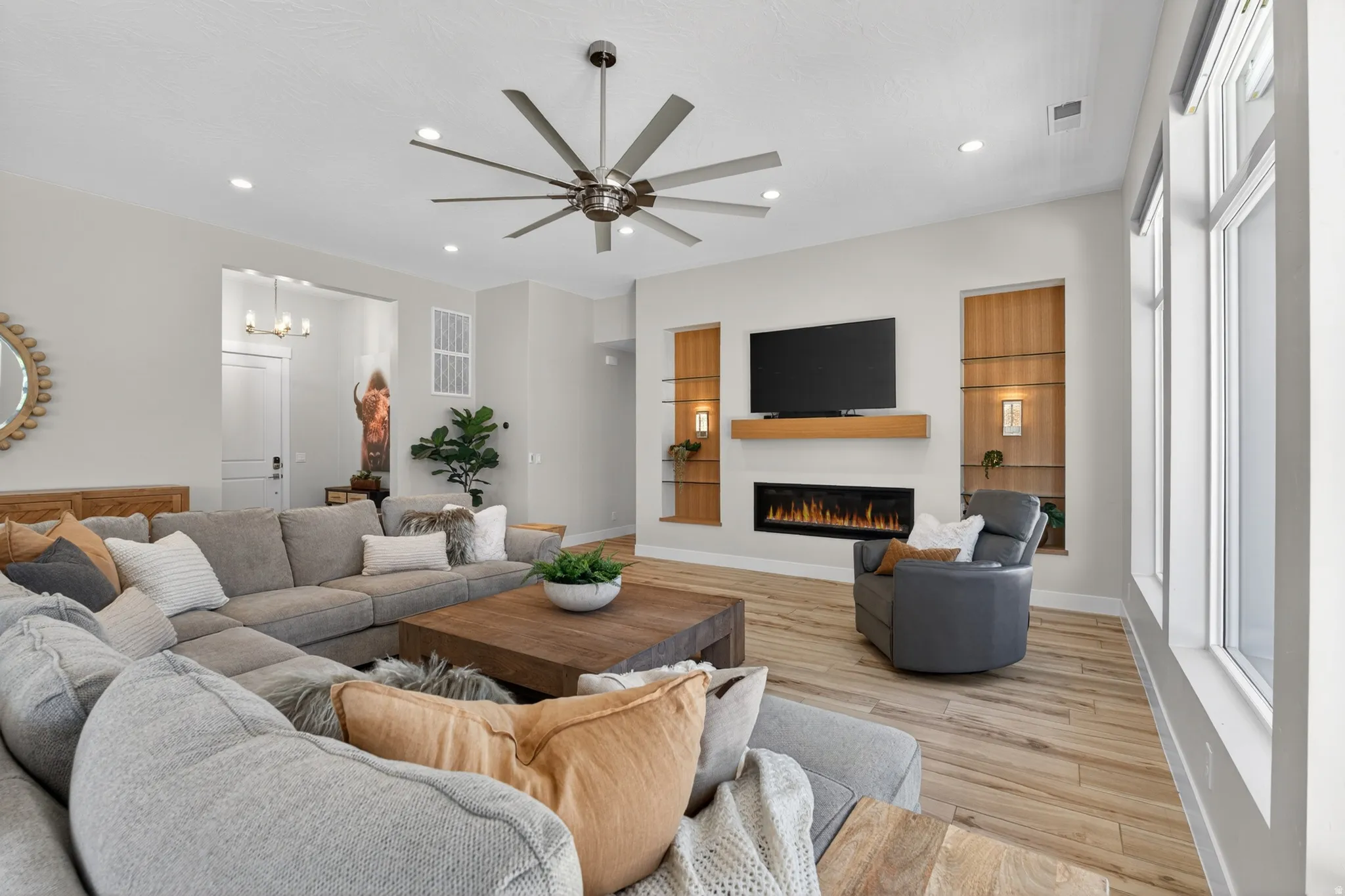 Spacious living area featuring a modern linear oversized fireplace with a wood mantel, built-in shelving with accent lighting, expansive windows, recessed lighting with glass shelving.
