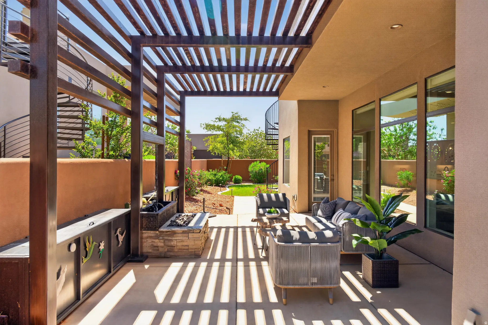 Spacious outdoor patio featuring a slatted pergola, integrated fire pit with stone surround, beautiful water fall and multiple large windows