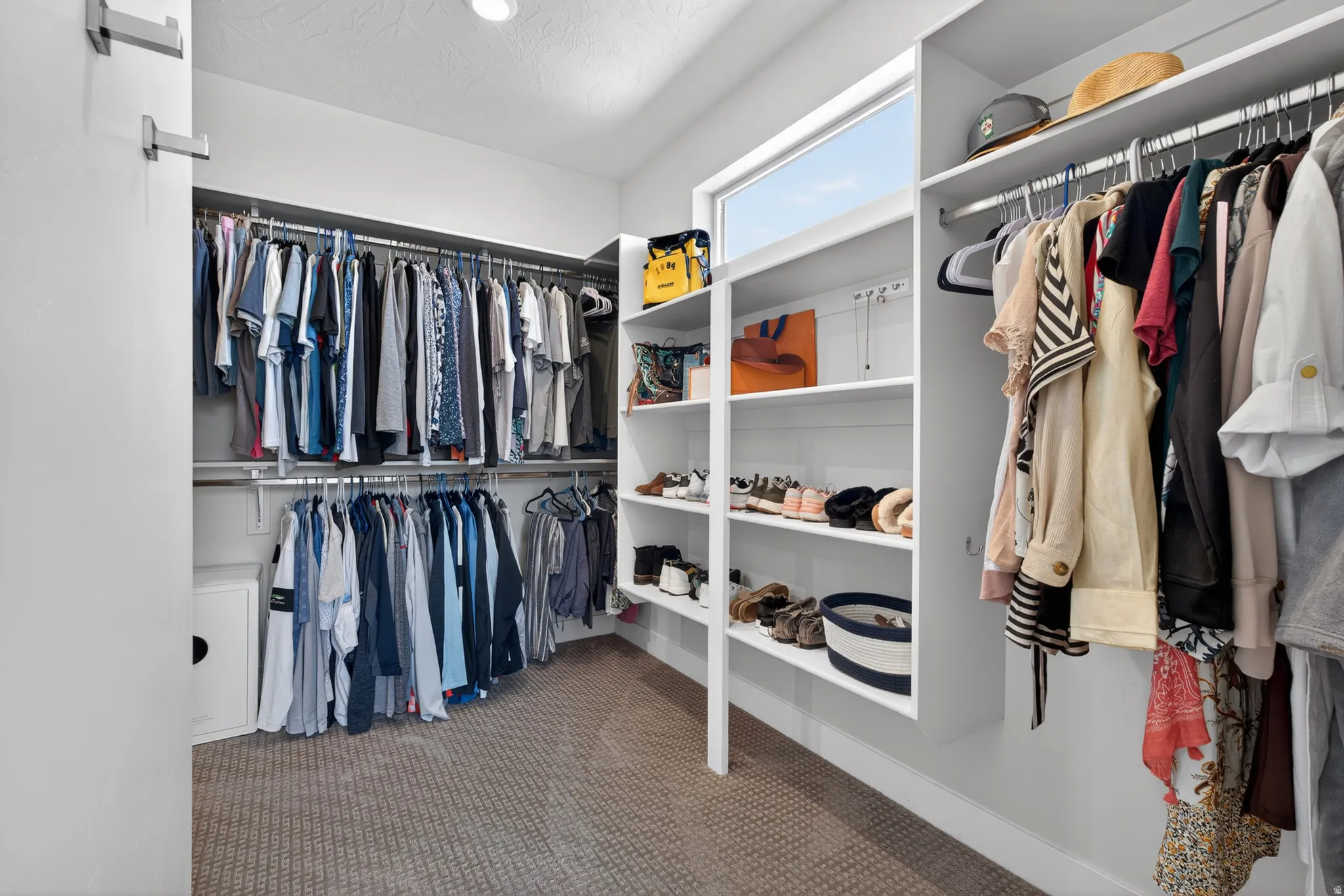 Spacious walk-in closet featuring white built-in shelving, chrome hanging rods, a high horizontal window, and patterned carpeting