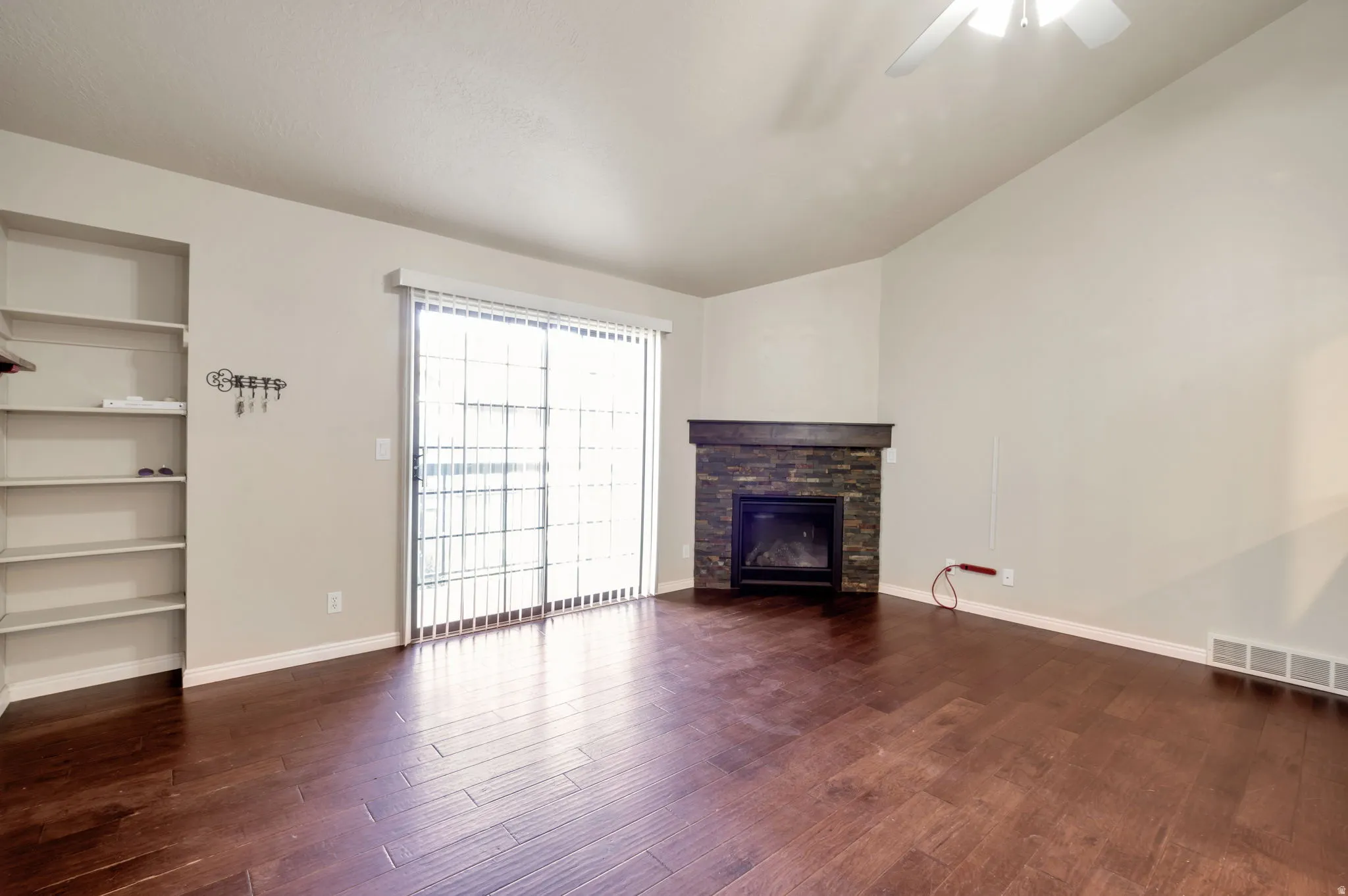 Spacious room featuring wood-finish flooring, a stacked stone fireplace with a dark wood mantel, built-in shelving, a sliding glass door, and a ceiling fan