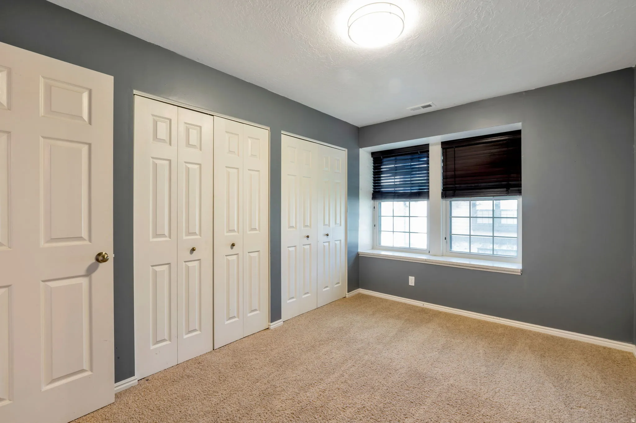 Bedroom featuring two panel doors, two bi-fold closet doors, two windows with dark blinds, and neutral carpet flooring