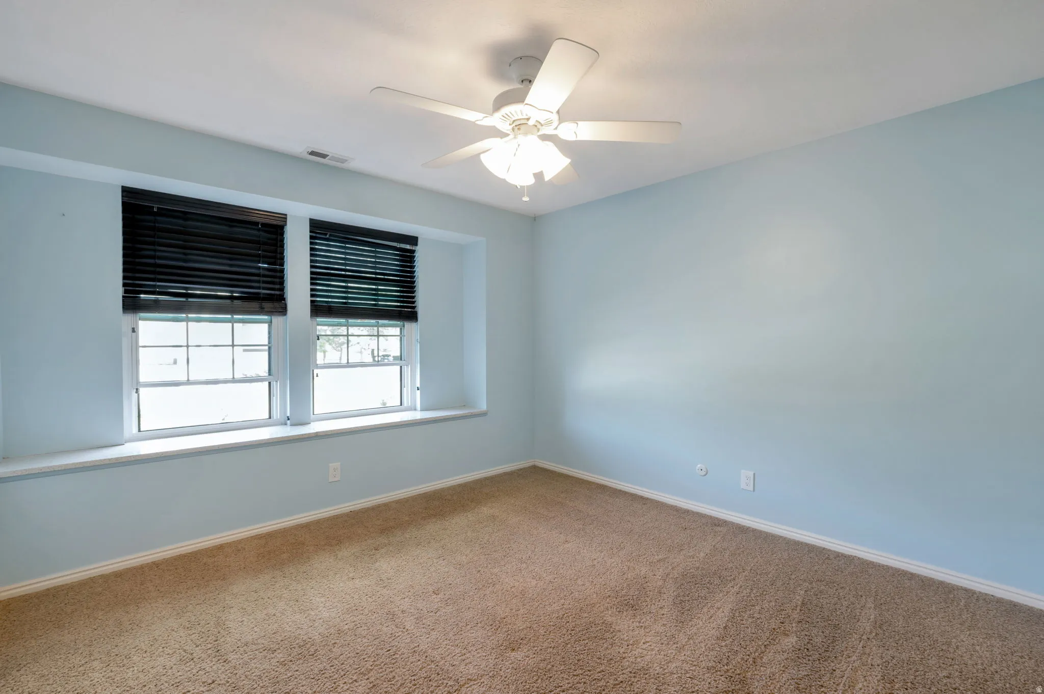 Carpeted room featuring light blue walls, two windows with dark blinds, a built-in window bench, and a ceiling fan with integrated lighting