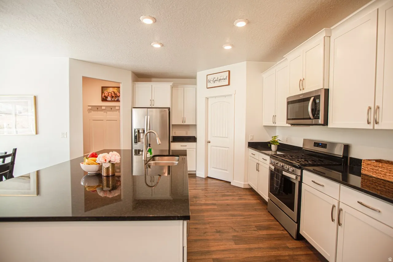 Modern kitchen featuring white cabinetry, dark countertops, stainless steel appliances, a kitchen island with an undermount sink, and wood-finish flooring