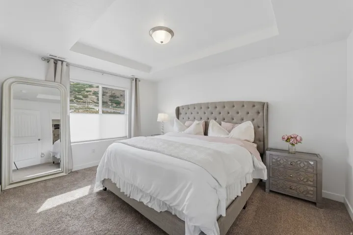 Bright master bedroom featuring a tufted upholstered headboard, wood-finish nightstand with drawer storage, recessed ceiling detail, and abundant natural light from dual windows