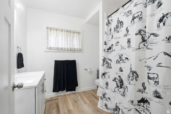 Bathroom featuring a white vanity with an integrated sink, wood-finish flooring, a window with sheer curtains, a toilet, and a shower area