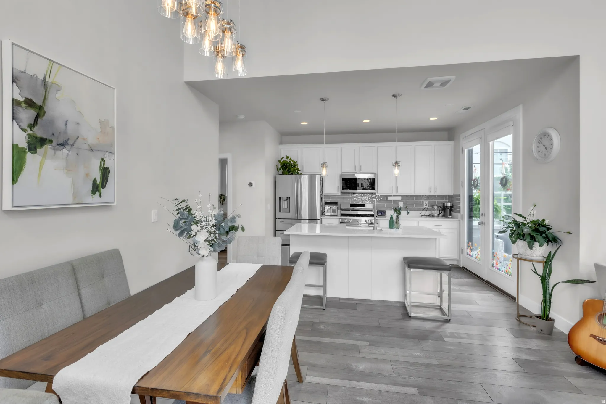 Open-concept living space featuring wood-finish flooring, a contemporary dining area with a modern chandelier, and a kitchen with white cabinetry and stainless steel appliances