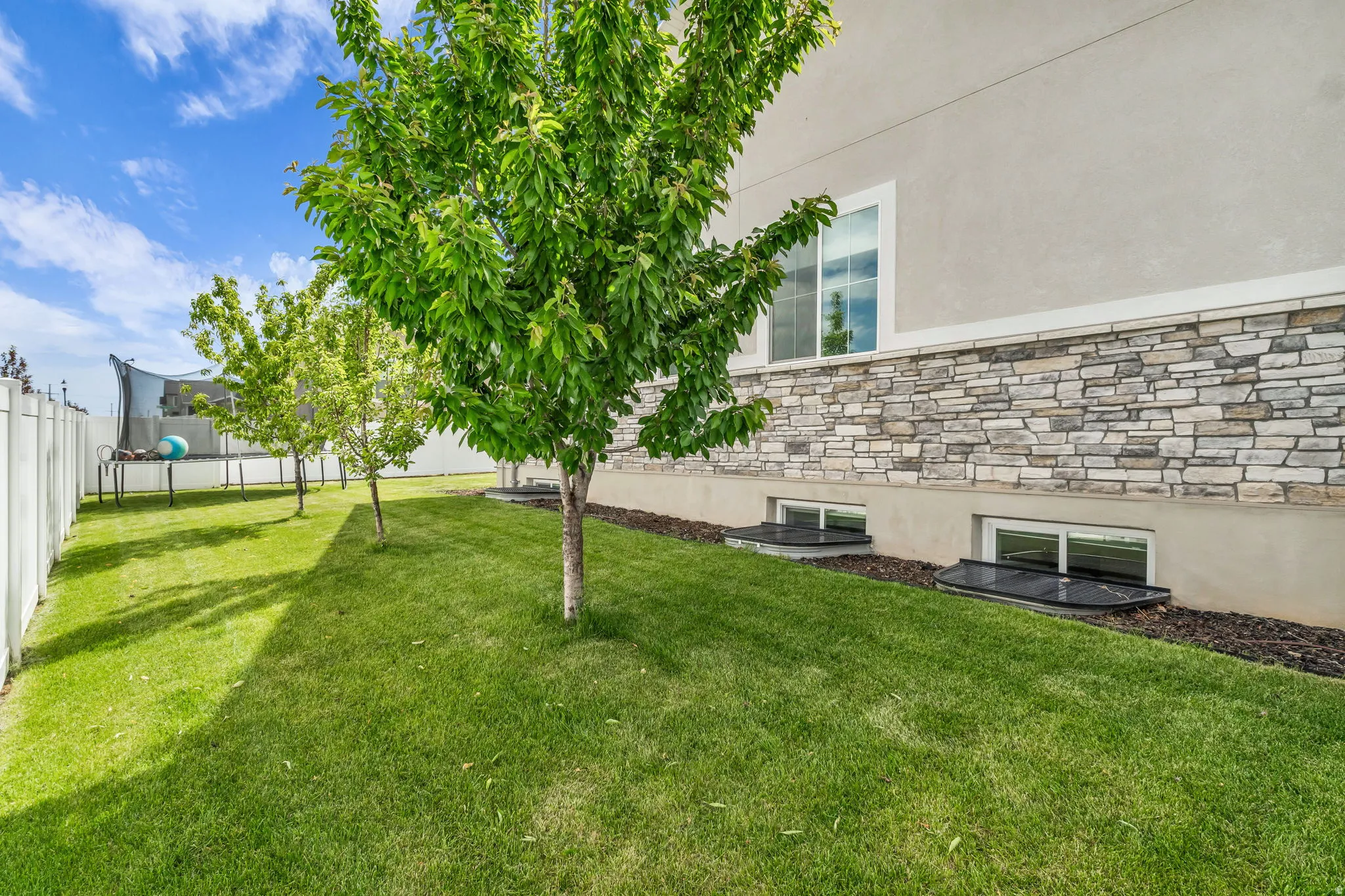 Manicured lawn with young trees, white privacy fencing, and a stucco exterior with stone veneer accents