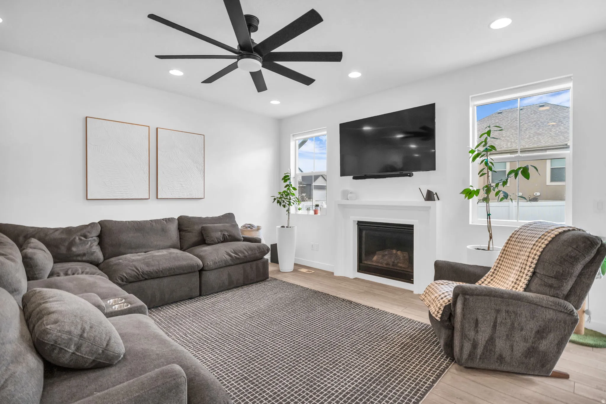 Living area featuring a modern black ceiling fan, recessed lighting, wood-finish flooring, a white fireplace with a dark insert, and multiple windows