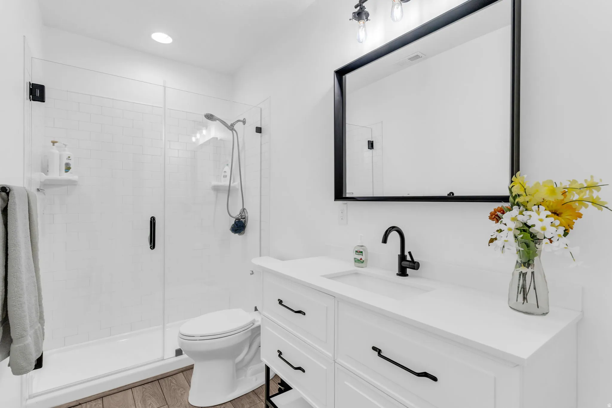 Modern bathroom featuring a frameless glass shower enclosure with white subway tile, a single vanity with an integrated sink and white countertop, black fixtures, and wood-finish flooring