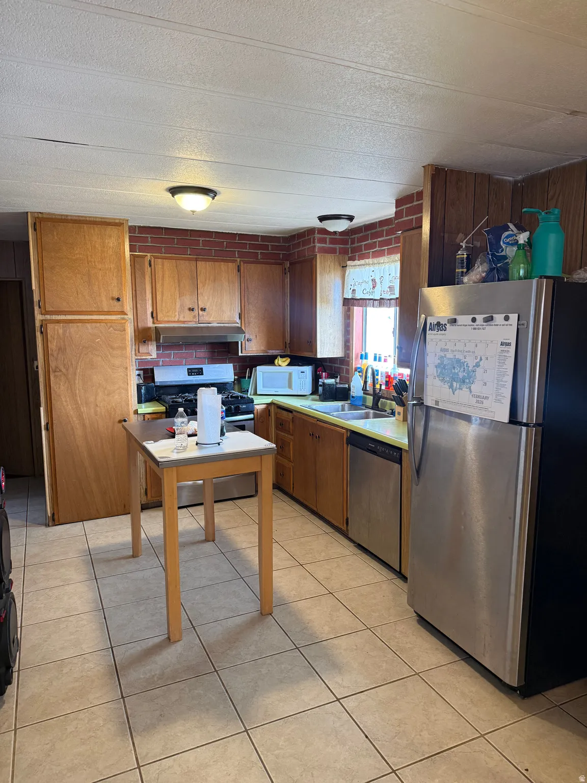 Kitchen featuring wood-finish cabinetry, tile flooring, brick-patterned wall accents, and stainless steel appliances