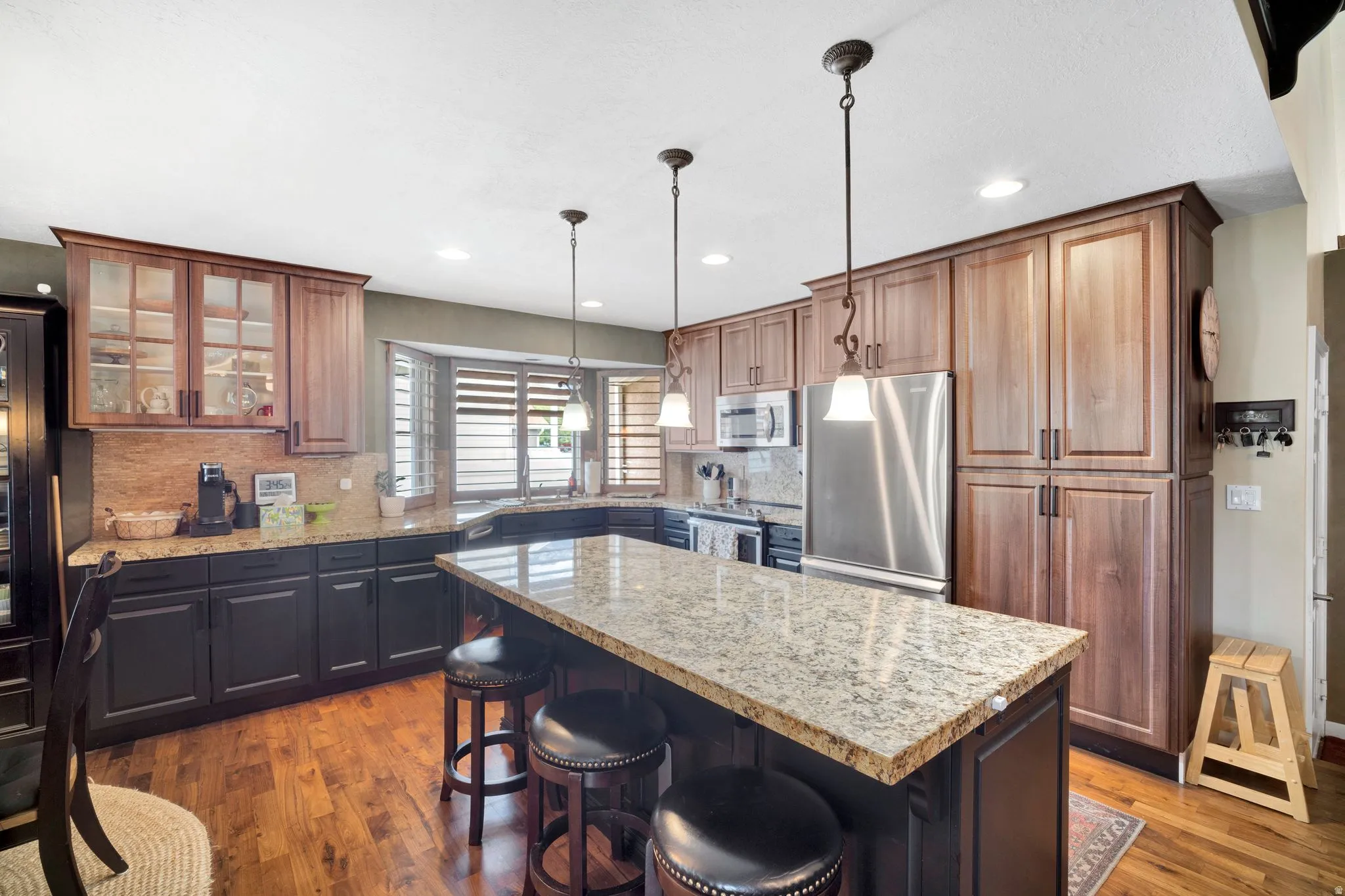 Modern kitchen featuring granite countertops, large island, pendant lighting, stainless steel appliances, walnut flooring, roll out shelving in pantry, and a bay window over sink.