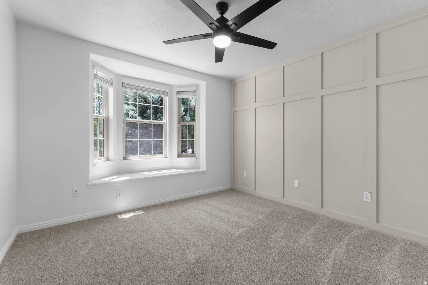 Room featuring a bay window, window seat, neutral carpet flooring, an accent wall with wainscoting, and a ceiling fan