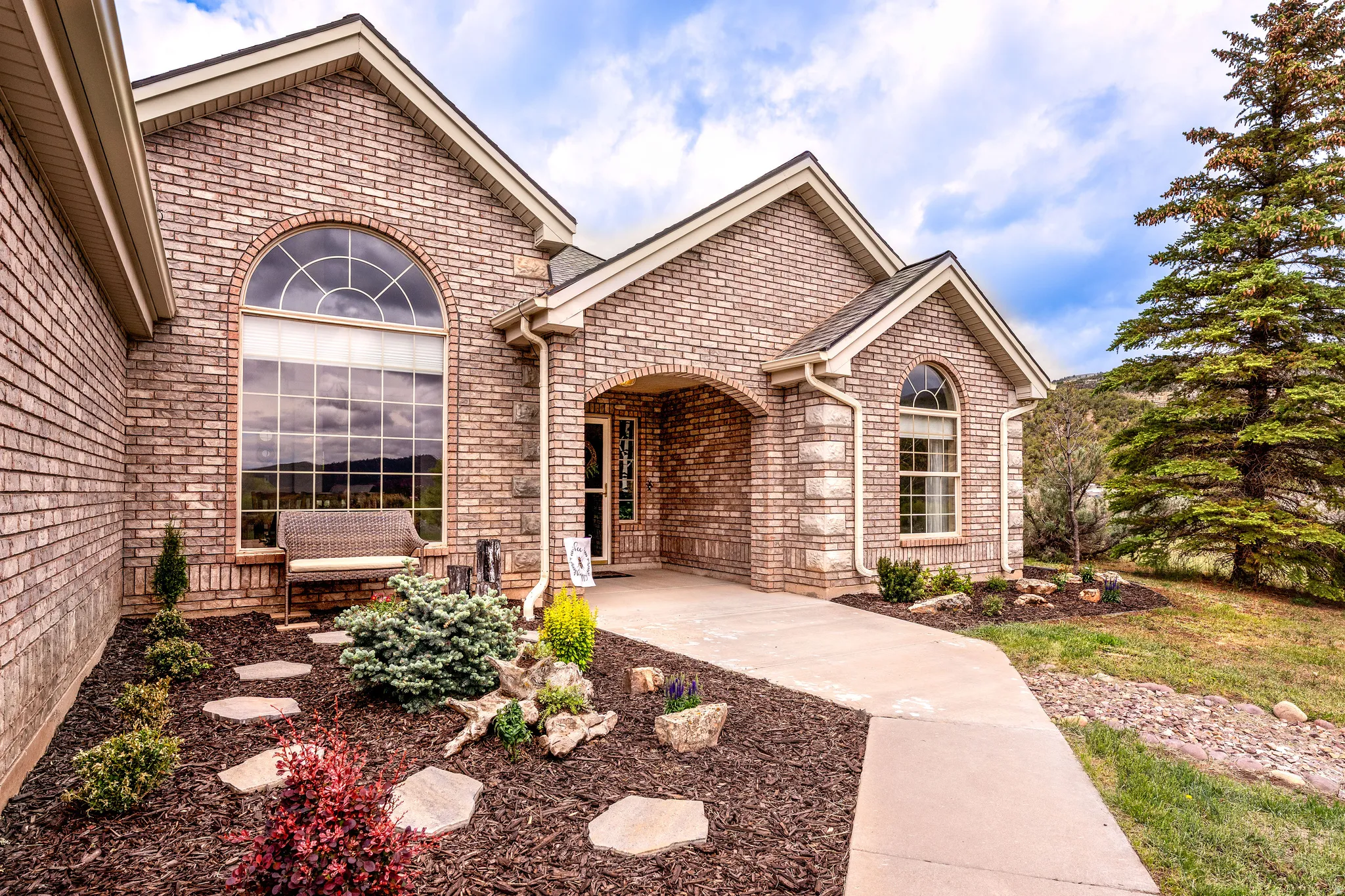 Brick exterior featuring an arched transom window, a prominent entry archway, a concrete pathway, landscaped garden beds with decorative rocks and mulch, and a mature evergreen tree