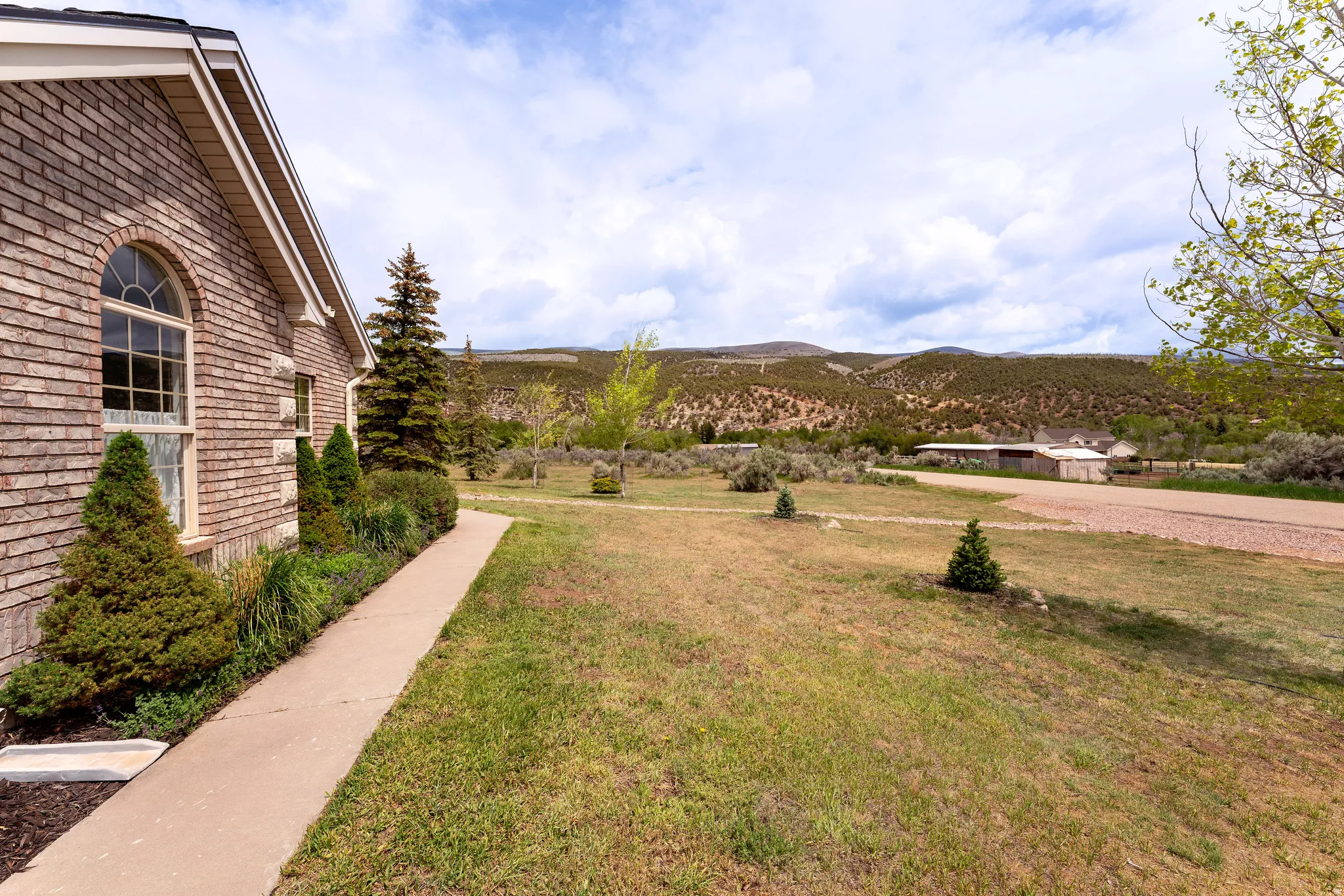 Brick exterior with arched window, concrete walkway, mature evergreen landscaping, and expansive yard with distant mountain backdrop