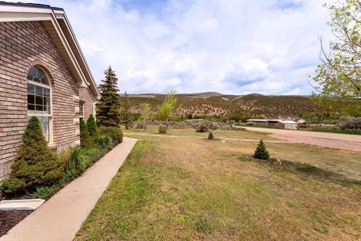 Brick exterior with arched window, concrete walkway, mature evergreen landscaping, and expansive yard with distant mountain backdrop