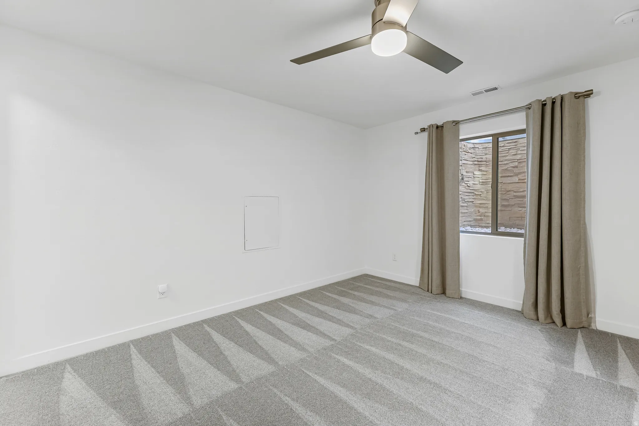 Basement bedroom with white walls, patterned gray carpet, and a window featuring stone exterior detailing
