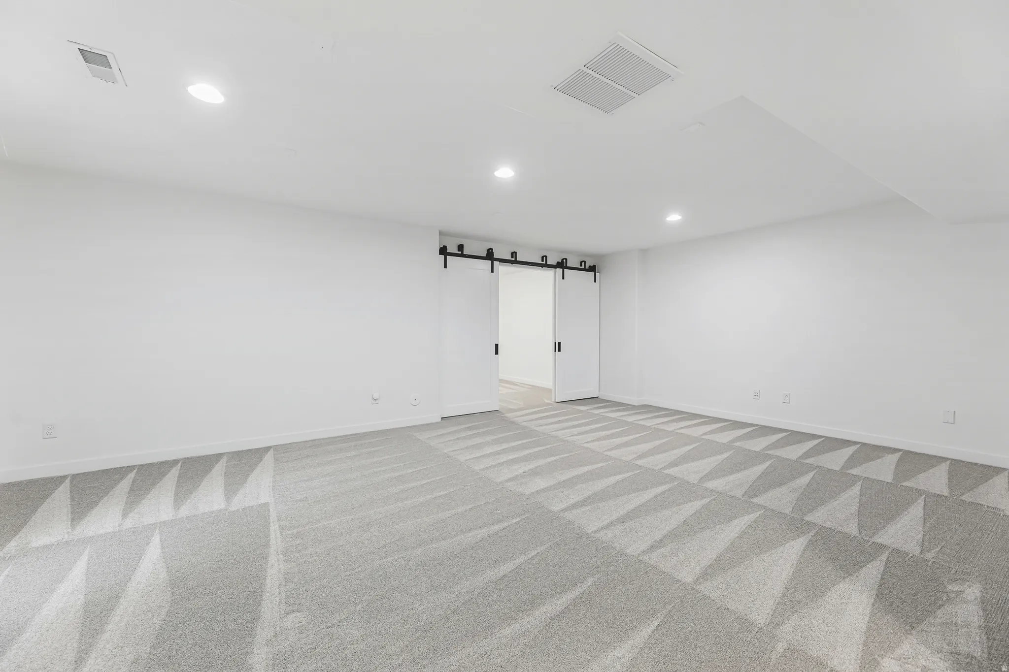 Spacious basement family room featuring patterned gray carpeting, white walls, recessed lighting, and a modern barn door entry