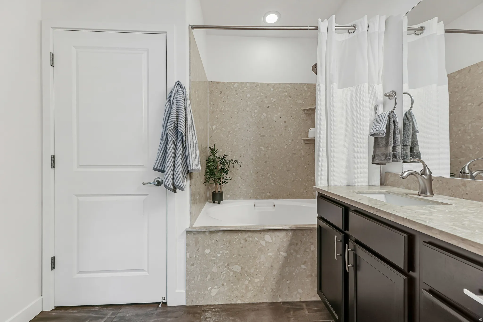 Primary bathroom featuring a built-in tub with speckled surround, a single vanity with dark cabinetry, a light-toned countertop, and a wall-mounted mirror