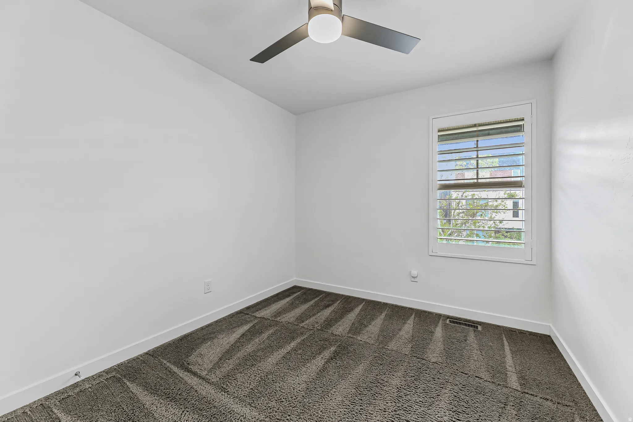 Bright bedroom featuring a window with white shutters, overhead ceiling fan with light, and textured carpeting